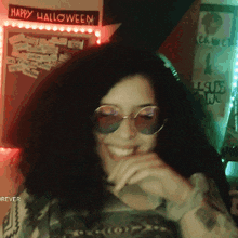 a woman wearing sunglasses smiles in front of a happy halloween sign