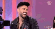 a man in a leather jacket is laughing while sitting on a stage in front of a camera .