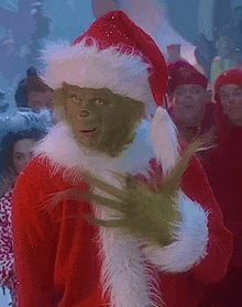 the grinch is wearing a santa hat and a red sweater and has green claws on his hands .