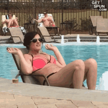 a woman in a bikini sits on a chair by a pool with a sign that says get spun.com