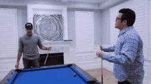 two men are playing pool in a living room and one has a hat on that says ' i love nyc '