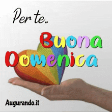 a person is holding a rainbow heart in their hand with the words `` buona domenica '' written on it .