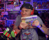 a man wearing headphones holds a comic book in front of a shelf with a sign that says maniac mansion