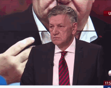 a man in a suit and tie stands in front of a screen that says ' 17:05 ' on it