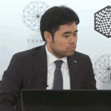 a man in a suit and tie is sitting in front of a laptop with the word tash on the wall behind him