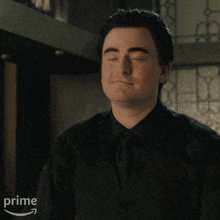 a man wearing a black shirt and a black vest is standing in front of a window with an amazon prime logo in the corner