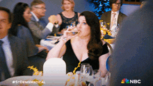 a woman sits at a table drinking from a glass with a nbc logo on the bottom