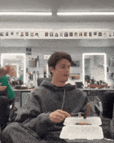 a man in a gray hoodie is eating a hot dog from a styrofoam box