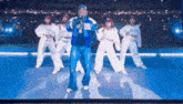 a man in a blue jacket is singing into a microphone while a group of dancers are dancing around him .