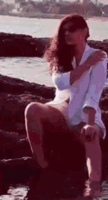 a woman in a white shirt and shorts is sitting on a rock near the water .
