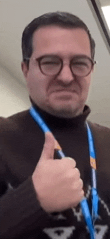 a man with glasses and a lanyard around his neck is giving a thumbs up .