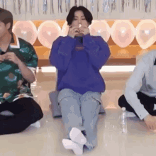 a group of people are sitting on the floor with balloons in the background . one of the men is wearing a blue sweatshirt .