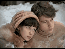 a boy and a girl are hugging in the snow . the girl is wearing a knitted hat .