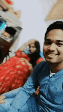 a man in a blue shirt smiles while a woman in a red dress sits behind him