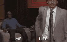 a man in a suit and tie is standing in front of a microphone while another man sits on a couch .