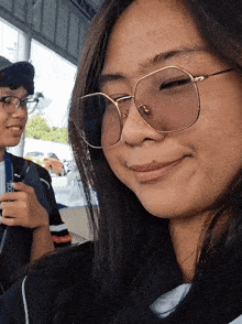 a woman wearing glasses looks at the camera with a man behind her