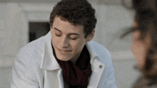 a young man wearing a white coat and scarf is smiling while talking to a woman .