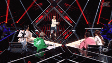 a group of people are sitting at a table in front of a stage that has a x on it