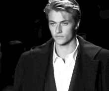 a black and white photo of a man in a suit and white shirt walking down a runway at a fashion show .