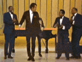 a group of men in suits and bow ties are dancing on a stage in front of a piano .