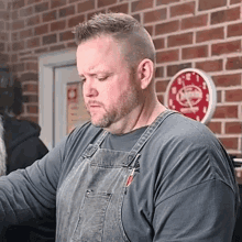 a man wearing overalls stands in front of a brick wall and a clock that says ' noodles '