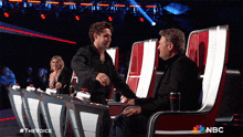 a man shakes hands with another man while sitting in front of a nbc logo