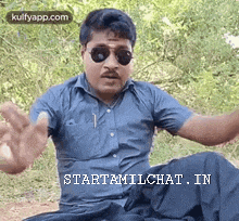 a man wearing sunglasses and a blue shirt is sitting on the ground with his arms outstretched .