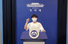 a man wearing a mask stands at a podium in front of a blue wall with korean writing on it
