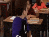 a boy in a purple shirt with the letter l on it is sitting at a desk in a classroom with other children .