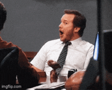 a man in a white shirt and tie is sitting at a table with his mouth open ..