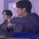 a man in a suit is sitting next to another man in a blue chair and holding his hand out .