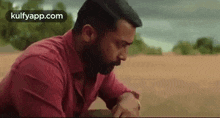 a man with a beard and a red shirt is sitting on the ground in a field .