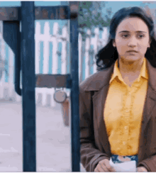 a woman in a yellow shirt and brown jacket stands in front of a gate
