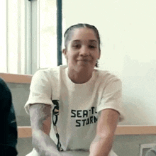 a woman wearing a white t-shirt with a tattoo on her arm is sitting in front of a window .