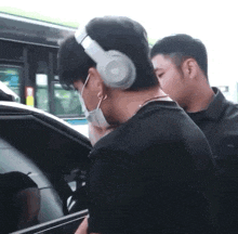 a man wearing headphones and a face mask is walking towards a car .