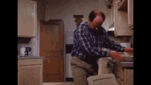 a man in a plaid shirt is standing in a kitchen holding a trash can and plates .