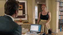 a man sitting at a desk with a laptop and a woman standing behind him saying i 'm going ..