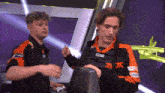 a man wearing a black and orange jacket with the letter f on it is standing next to another man