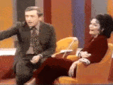 a man in a suit and tie is sitting next to a woman in a red dress who is sitting in a chair .