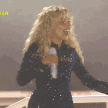 a woman with blonde curly hair is singing into a microphone in front of a yellow sign that says ter