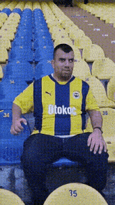 a man wearing a yellow and blue shirt that says otokac sits in a stadium
