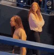 two women are standing next to each other in a stadium . one of the women has braids in her hair .