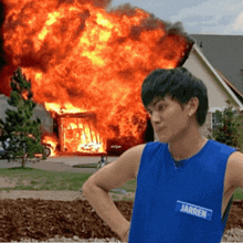 a man wearing a blue tank top with jarren on it stands in front of a burning house