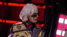 a woman wearing sunglasses and a wrestling belt is standing in a dark room .