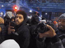 a man in a black hoodie holds a camera in front of a crowd of people