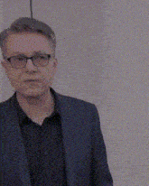 a man wearing a suit and glasses is standing in front of a white wall .