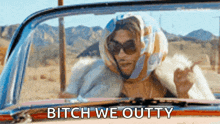 a man wearing sunglasses and a scarf on his head is driving a car and says bitch we outty .
