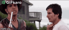 two men are standing next to each other in front of a house . one of the men is holding a gun in his mouth .