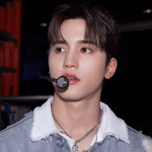 a young man wearing a denim jacket and a necklace holds a microphone in his mouth