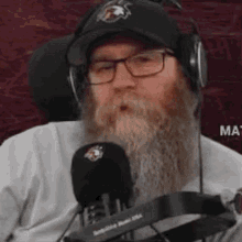 a man with a beard and glasses is wearing headphones and a hat while sitting in front of a microphone .